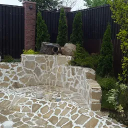 Kamienna konstrukcja ogrodowa z białymi fugami, otoczona zielenią i elementami dekoracyjnymi, w tle ciemne ogrodzenie.