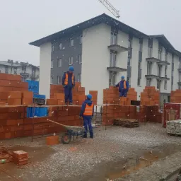 Budowa wielorodzinnego domu z czerwonej cegły ceramicznej w trakcie zimy, robotnicy w kaskach i kamizelkach odblaskowych murowują ściany, widoczny dźwig budowlany w tle.