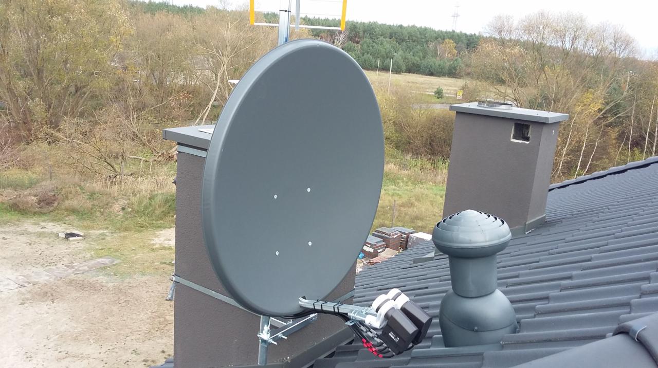 Szara antena satelitarna zamontowana na kominie na dachu budynku, widoczne kable i uchwyty montażowe, tło stanowią drzewa i łąka.