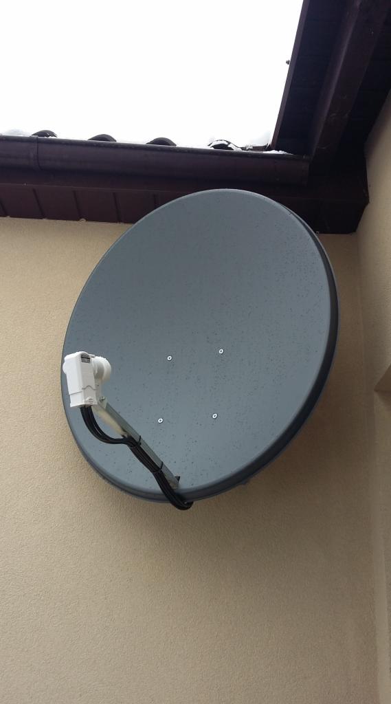 Szara antena satelitarna zamontowana na beżowej elewacji budynku, widoczne kable i fragment dachu z brązową rynną.