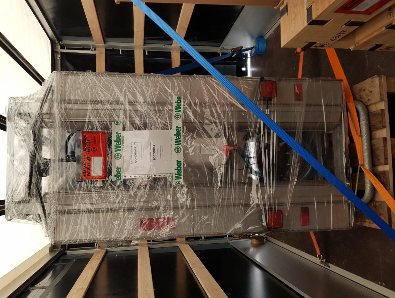 Zapakowany i zabezpieczony pasami transportowymi metalowy element z logo Weber, umieszczony w przestrzeni ładunkowej pojazdu na drewnianej palecie.