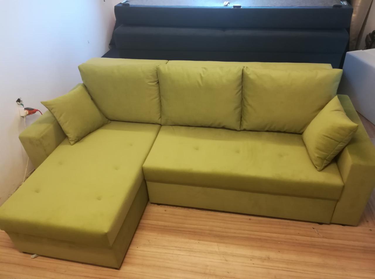 Limonkowy narożnik z welurową tapicerką, widoczne pikowania na siedzisku i poduszce, na podłodze panele drewniane, w tle ciemna kanapa.