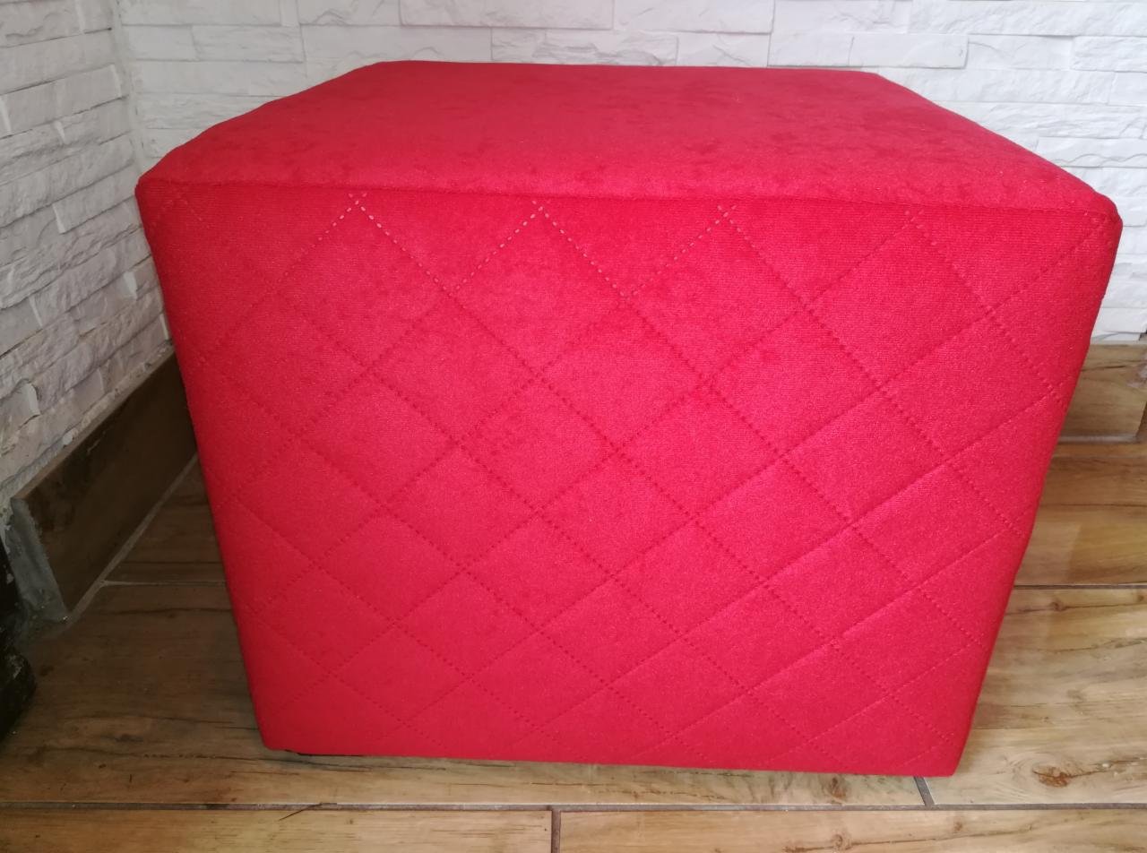 Czerwony, tapicerowany puf w kształcie sześcianu z pikowaniem w romby, stojący na drewnianej podłodze przy białej ścianie z imitacją cegły.