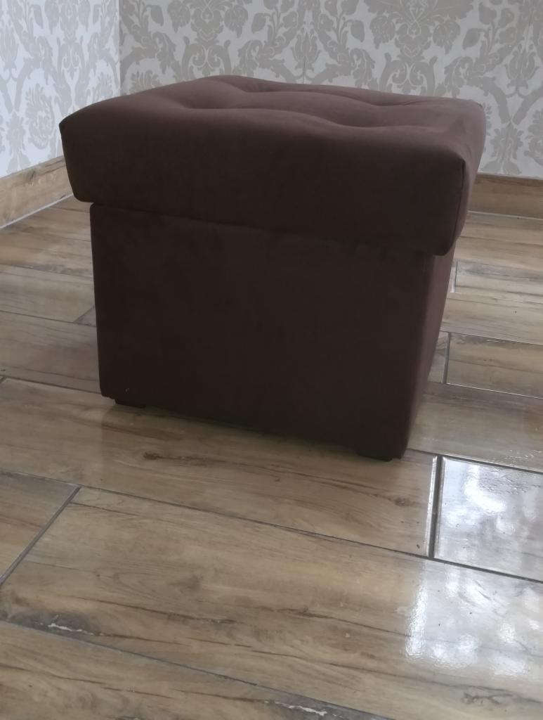 Ciemnobrązowy, tapicerowany puf z guzikami na siedzisku, stojący na drewnopodobnych płytkach w pomieszczeniu z tapetą w delikatny wzór.