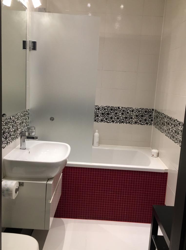 Nowoczesna łazienka z białą umywalką na szafce, kabiną prysznicową z matowego szkła i wanną obudowaną czerwoną mozaiką, akcentowana czarnym, kwiatowym wzorem na płytkach.