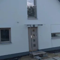 Elewacja domu jednorodzinnego z jasną strukturą tynku, widoczny fragment palety przed wejściem i okno z balustradą na piętrze.