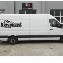 Biały van z logo firmy dekarskiej 'Kowalczyk Dachy' zaparkowany przed budynkiem. Widok z boku, logo wyraźnie widoczne. Prezentacja firmy.