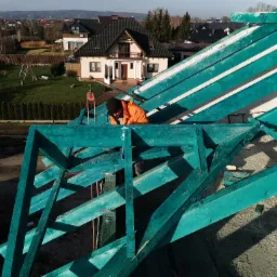 Pracownik w pomarańczowej kurtce montuje pomalowaną na turkusowo drewnianą konstrukcję dachu na tle domów jednorodzinnych.