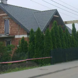 Niedokończony dom murowany z szarym dachem, widoczna cegła, ogrodzony szpalerem tui, taśma ostrzegawcza przy ziemi.