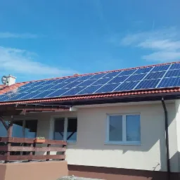 Dom jednorodzinny z zainstalowanymi panelami słonecznymi na dachu z czerwonej dachówki, widoczne okna i drewniana weranda, błękitne niebo z chmurami w tle.