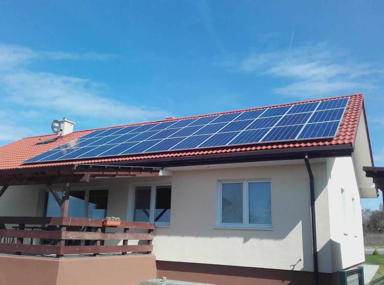 Dom jednorodzinny z zainstalowanymi panelami słonecznymi na dachu z czerwonej dachówki, widoczne okna i drewniana weranda, błękitne niebo z chmurami w tle.