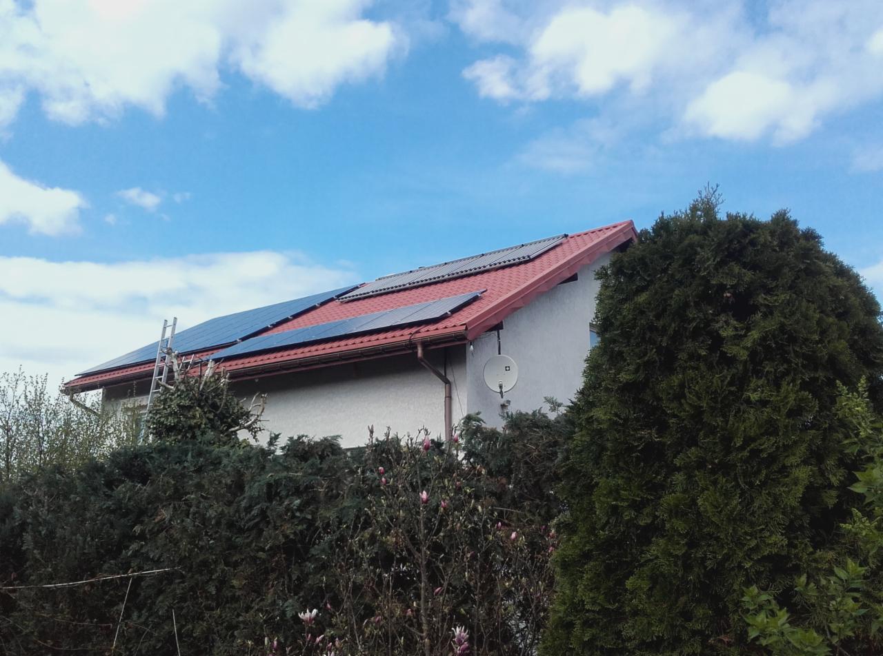 Instalacja fotowoltaiczna na dachu domu jednorodzinnego z czerwoną dachówką, widoczna drabina oparta o dach, antena satelitarna na ścianie, częściowo zasłonięta przez zieleń.