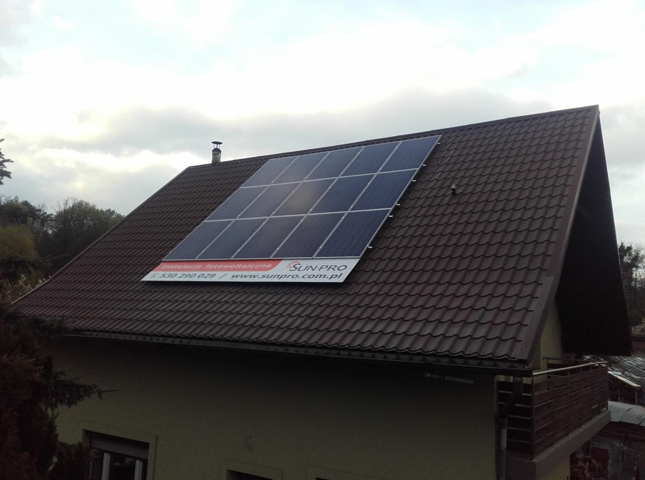 Panele słoneczne zamontowane na brązowym dachu skośnym domu jednorodzinnego z widocznym banerem reklamowym firmy instalującej.