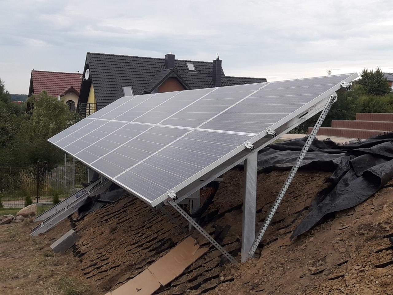 Panel słoneczny na metalowej konstrukcji, zamontowany na zboczu ziemi, w tle domy jednorodzinne.