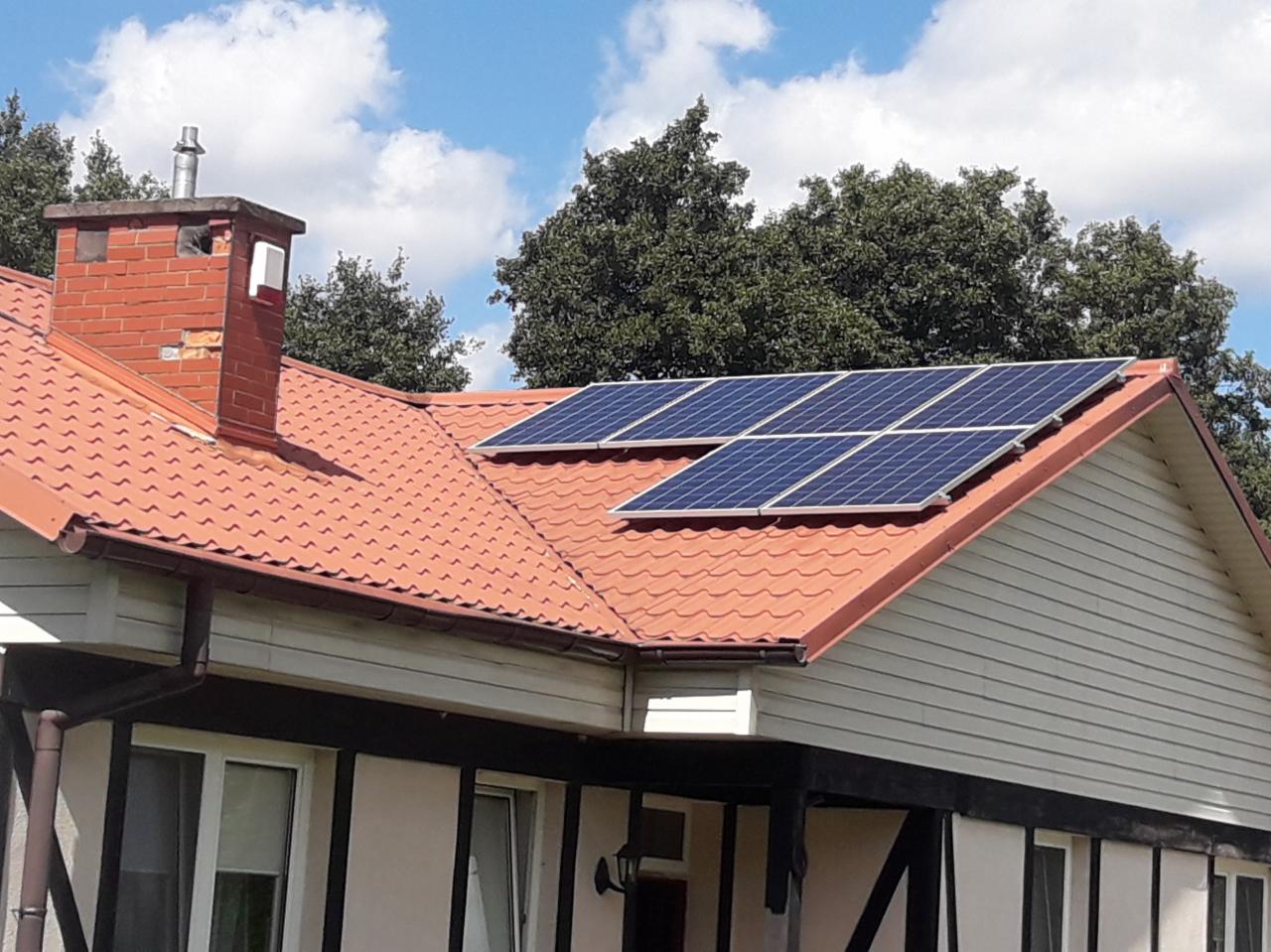 Instalacja paneli słonecznych na dachu domu jednorodzinnego z czerwoną dachówką, widoczny komin z cegły i fragment elewacji w kolorze jasnego beżu.