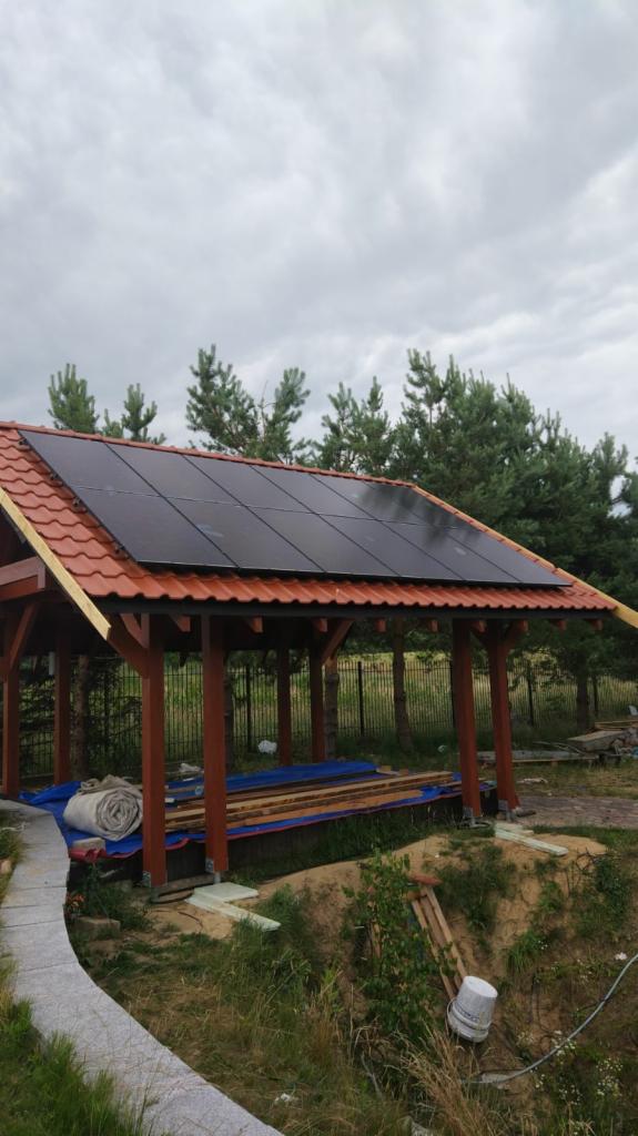 Altana z czerwoną dachówką, na której zamontowano czarne panele fotowoltaiczne, w tle iglaste drzewa i pochmurne niebo.