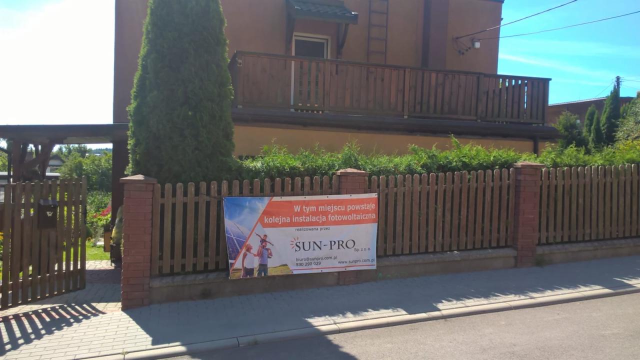 Baner reklamowy firmy SUN-PRO zapowiadający instalację na drewnianym ogrodzeniu z ceglanymi słupkami, w tle dom z balkonem i zielenią.