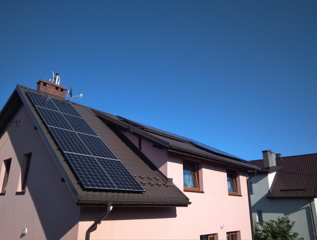 Panele słoneczne na dachu domu jednorodzinnego z brązową dachówką, widoczne kominy i antena satelitarna, błękitne niebo w tle.