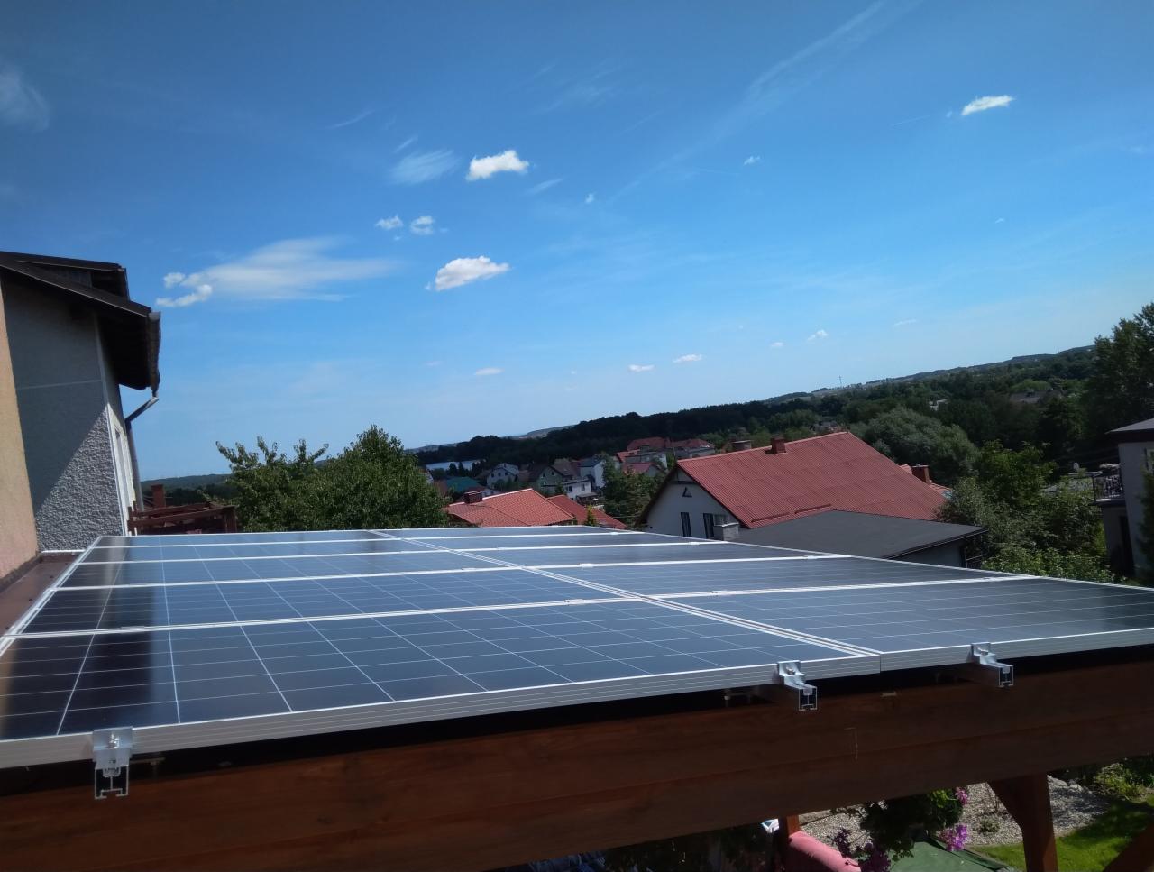 Instalacja paneli słonecznych na drewnianej konstrukcji dachu z widokiem na zabudowę jednorodzinną i błękitne niebo z lekkimi obłokami.