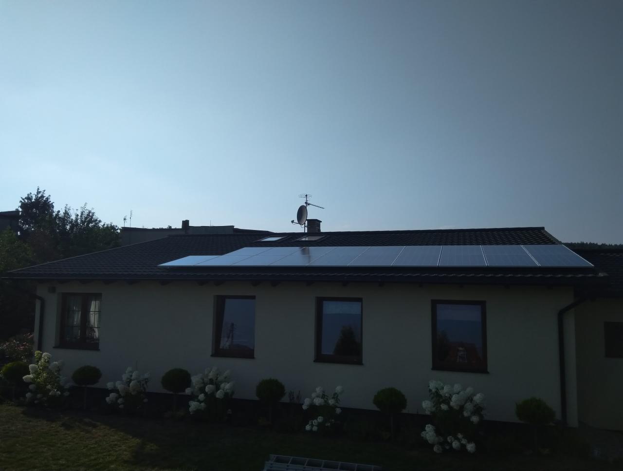 Panele słoneczne zamontowane na dachu domu jednorodzinnego z anteną satelitarną, widok z poziomu ogrodu z kwitnącymi krzewami.