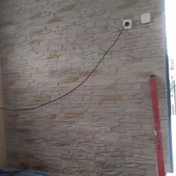 Ściana wykończona panelami imitującymi kamień, widoczne gniazdka elektryczne, poziomica pionowa przy ścianie, zabezpieczona podłoga folią i stos paneli pod ścianą.