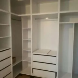 Biała, narożna garderoba z szufladami i półkami, oświetlona listwą LED, z widocznym fragmentem drewnianej podłogi.