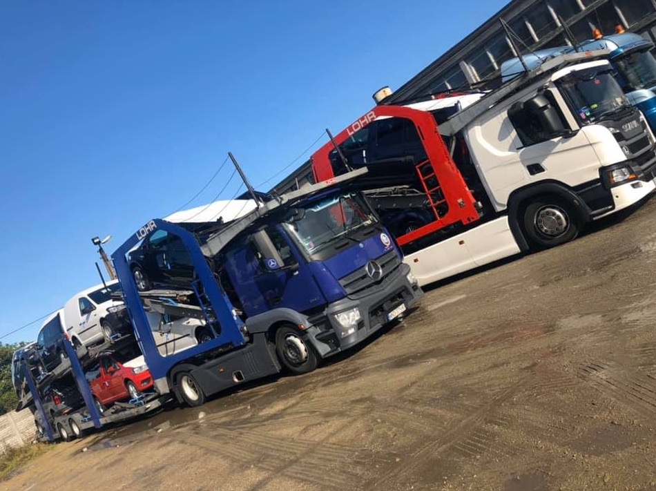 Dwa samochody ciężarowe typu laweta, jeden niebieski Mercedes i drugi biały Scania, załadowane samochodami osobowymi, stoją na placu w Pleszewie, gotowe do drogi.