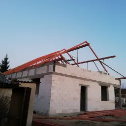 Wznoszenie ścian parterowego domu z betonu komórkowego, widoczne drewniane elementy konstrukcji dachu, w tle budynek gospodarczy i drzewa.