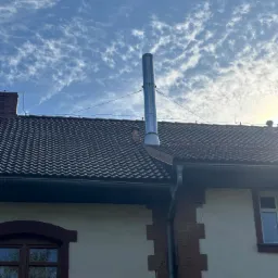 Błyszczący, metalowy komin wentylacyjny zamontowany na ciemnobrązowym dachu z dachówki ceramicznej, na tle błękitnego nieba z małymi, białymi obłokami.