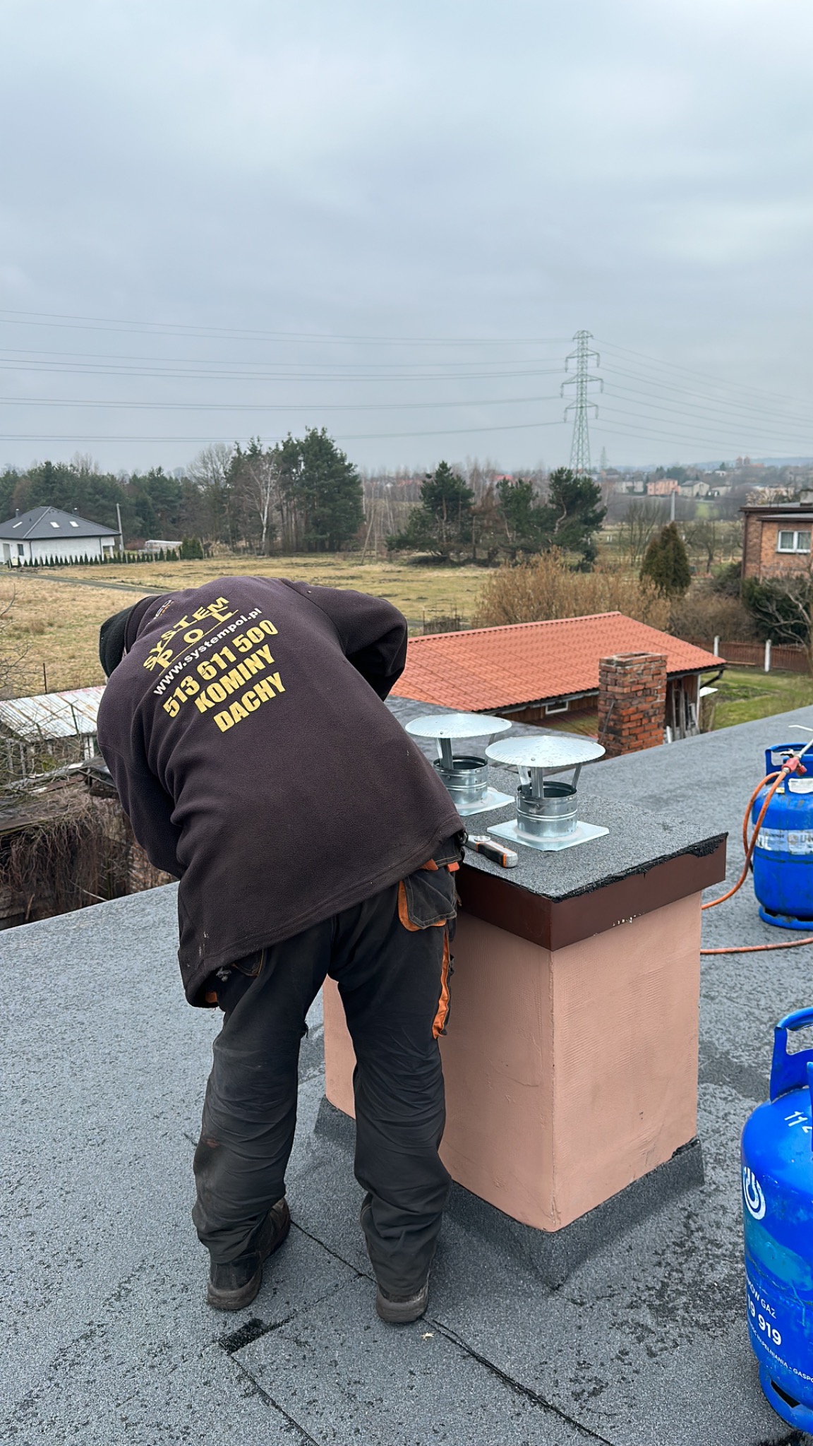 Pracownik w firmowej bluzie z logo montuje metalowe nasady kominowe na dachu, w tle zabudowa jednorodzinna i słupy wysokiego napięcia.