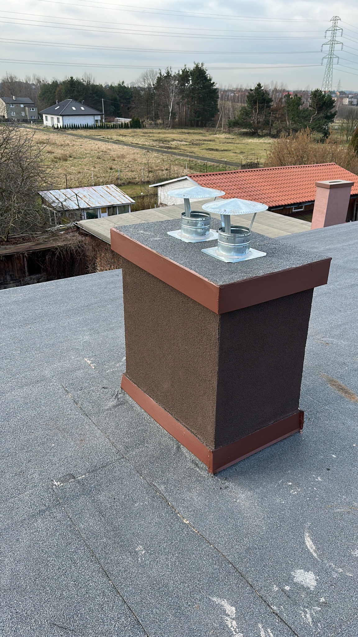 Widok z góry na brązowy komin z dwoma metalowymi nasadami na dachu pokrytym papą, w tle zabudowa jednorodzinna i linia wysokiego napięcia.