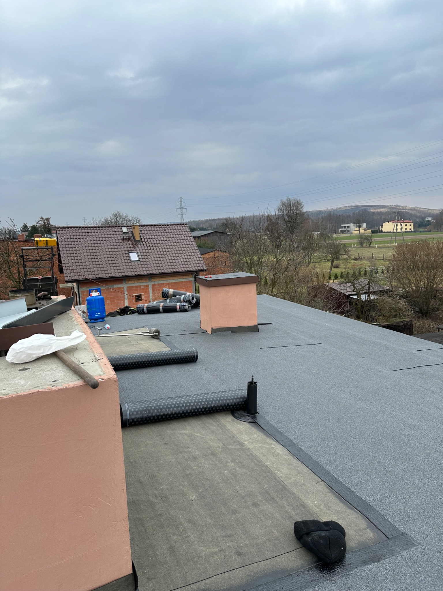 Widok z góry na płaski dach pokryty papą termozgrzewalną, z widocznymi rolkami papy, butlą gazową, kominem oraz fragmentem innego dachu w tle, na tle pochmurnego nieba.
