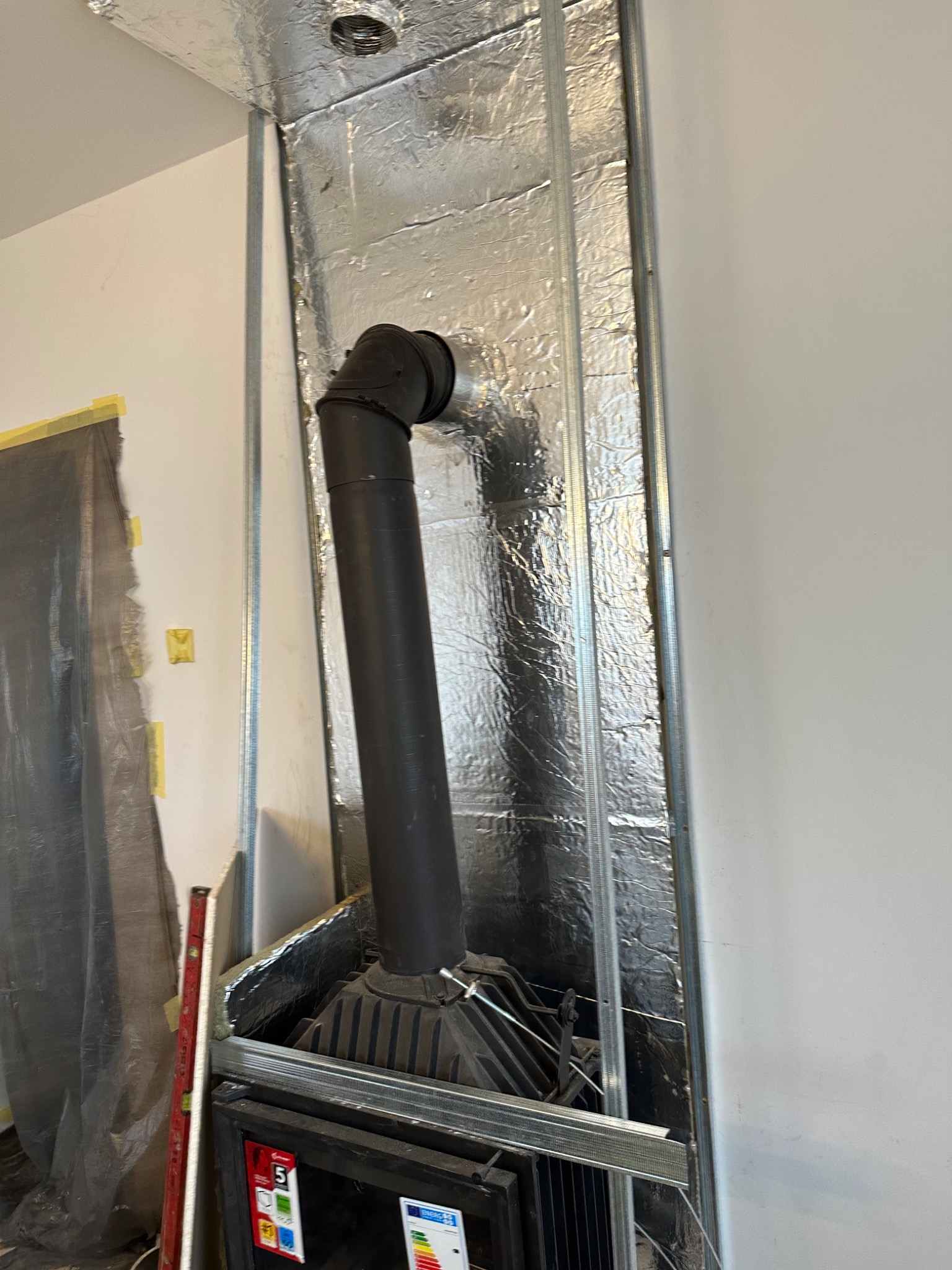 Wnętrze kominka w trakcie montażu, widoczne izolacja aluminiowa, stalowa konstrukcja, czarny przewód kominowy i poziomica oparta o ścianę.