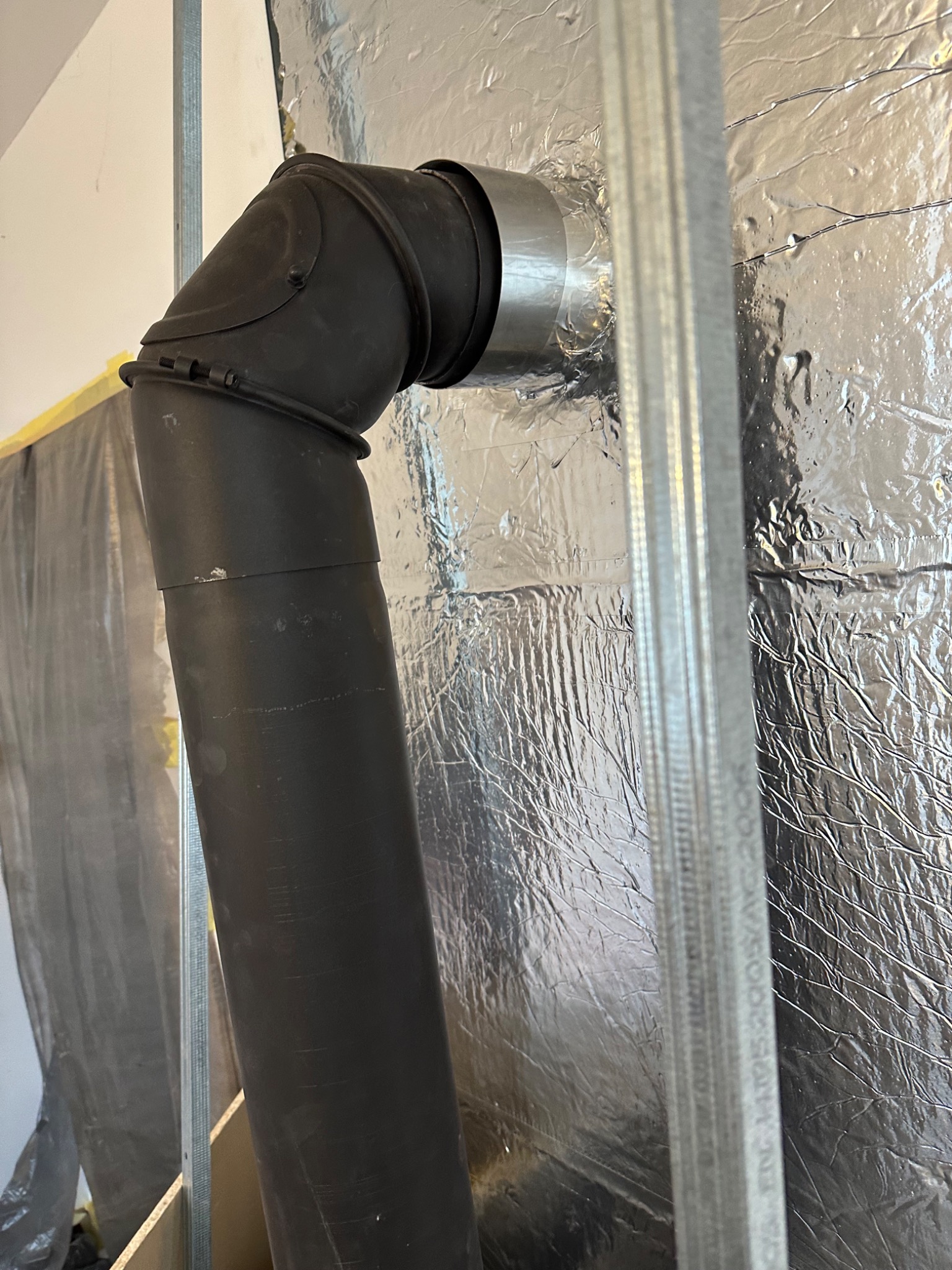 Czarny, metalowy przewód kominowy, połączony srebrną taśmą, przechodzi przez ścianę z folii aluminiowej, w tle widoczna konstrukcja szkieletowa.