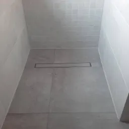 Szara kabina prysznicowa z odpływem liniowym w podłodze, wykończona płytkami w odcieniach szarości, widoczna słuchawka prysznicowa.