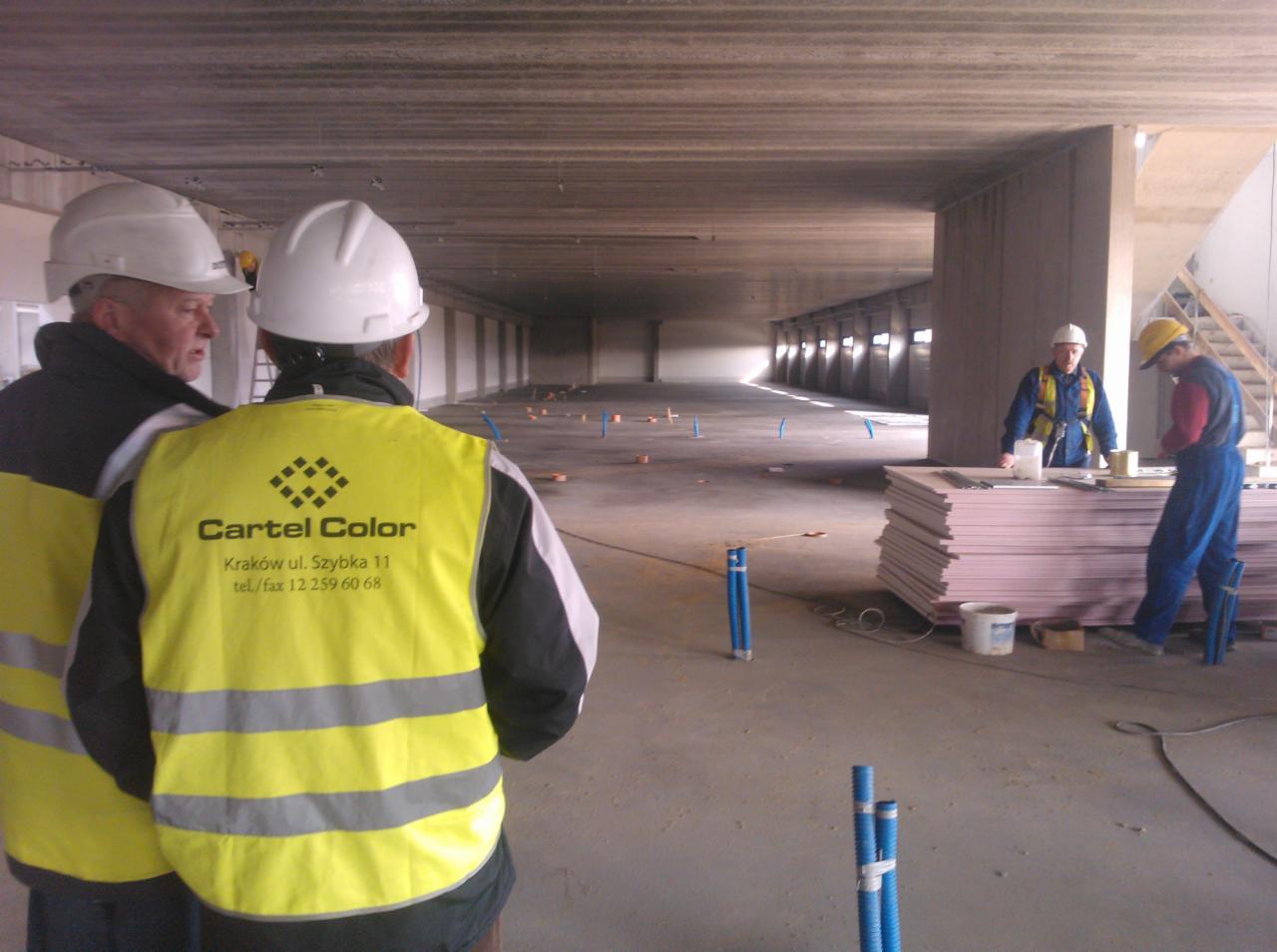 Dwóch mężczyzn w kaskach i kamizelkach odblaskowych z logo Cartel Color obserwuje prace wykończeniowe w surowym, betonowym wnętrzu hali. W tle pracownicy układają płyty izolacyjne.