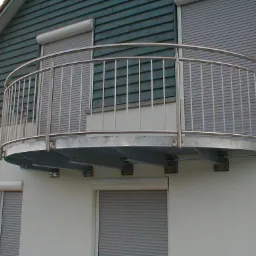 Zaokrąglony balkon z metalową konstrukcją i pionowymi prętami, zamontowany na białej elewacji budynku z szarymi roletami.