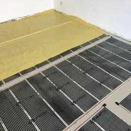 Rozłożona mata grzewcza na podłodze w trakcie instalacji, częściowo przykryta żółtą folią ochronną przy ścianie.