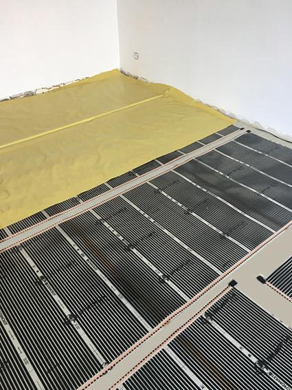 Rozłożona mata grzewcza na podłodze w trakcie instalacji, częściowo przykryta żółtą folią ochronną przy ścianie.
