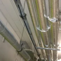 Metalowe rury z izolacją termiczną zamontowane pod sufitem w budynku, widoczne elementy mocujące i czarna osłona kabla.