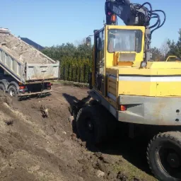 Żółta koparka kołowa Akerman i wywrotka z ładunkiem ziemi na placu budowy, widoczne drzewa w tle, słoneczny dzień.