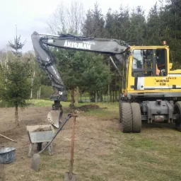 Żółta koparka Volvo Åkerman H5M z łyżką do sadzenia drzew, obok taczka z ziemią, szpadel i wiadro, na tle małych drzewek i lasu.