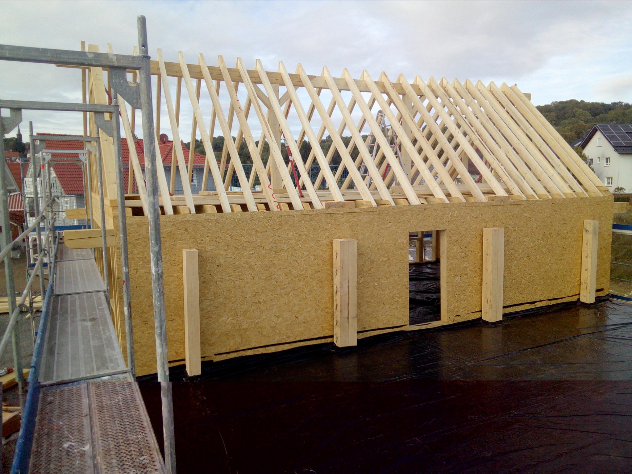 Surowa konstrukcja domu w trakcie budowy: ściany z płyt OSB, drewniany szkielet dachu, rusztowanie z boku, czarna folia hydroizolacyjna na podłożu.