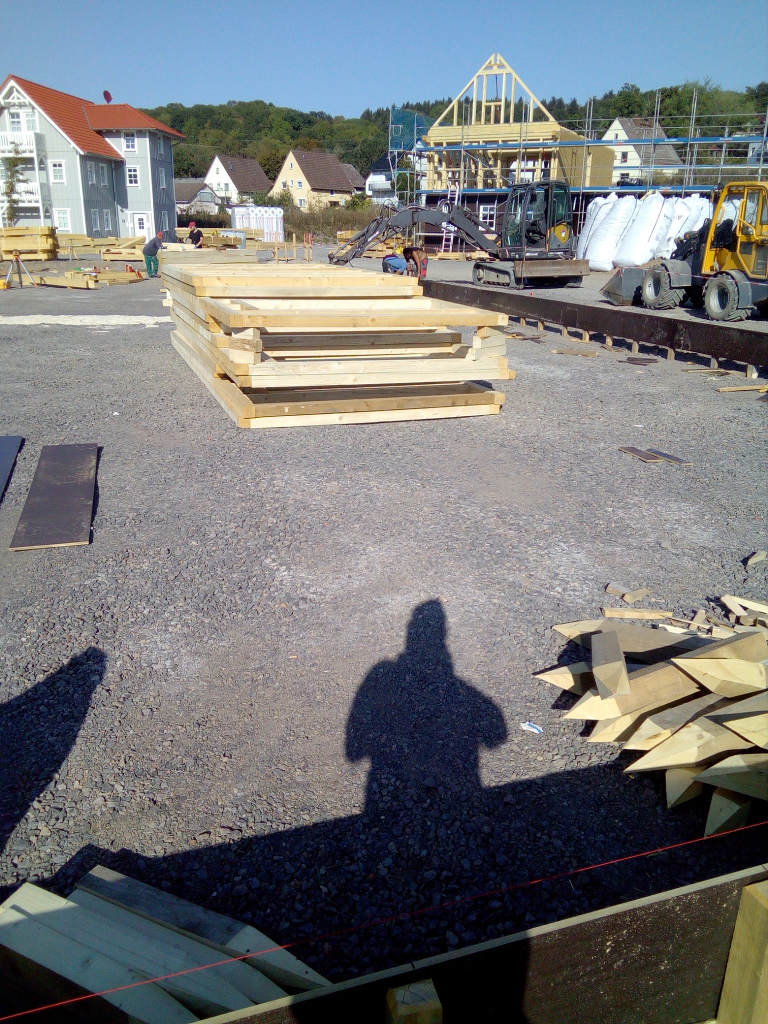 Stos drewnianych ram okiennych ułożony na żwirowym placu budowy, w tle szkielet konstrukcji domu i maszyny budowlane, cień sylwetki fotografa na pierwszym planie.