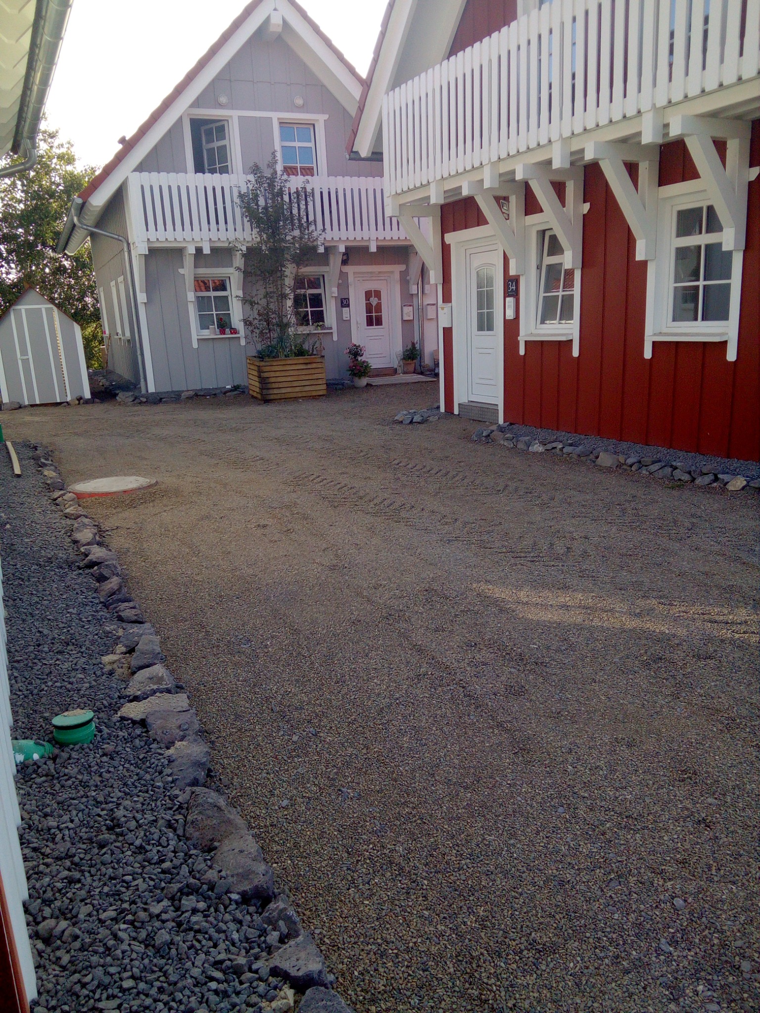 Dwa domy jednorodzinne, jeden szary z białymi detalami i balkonem, drugi czerwony z białymi detalami, oba z numerami domów, widoczne z perspektywy podwórza wyłożonego żwirem, z widocznymi śladami...
