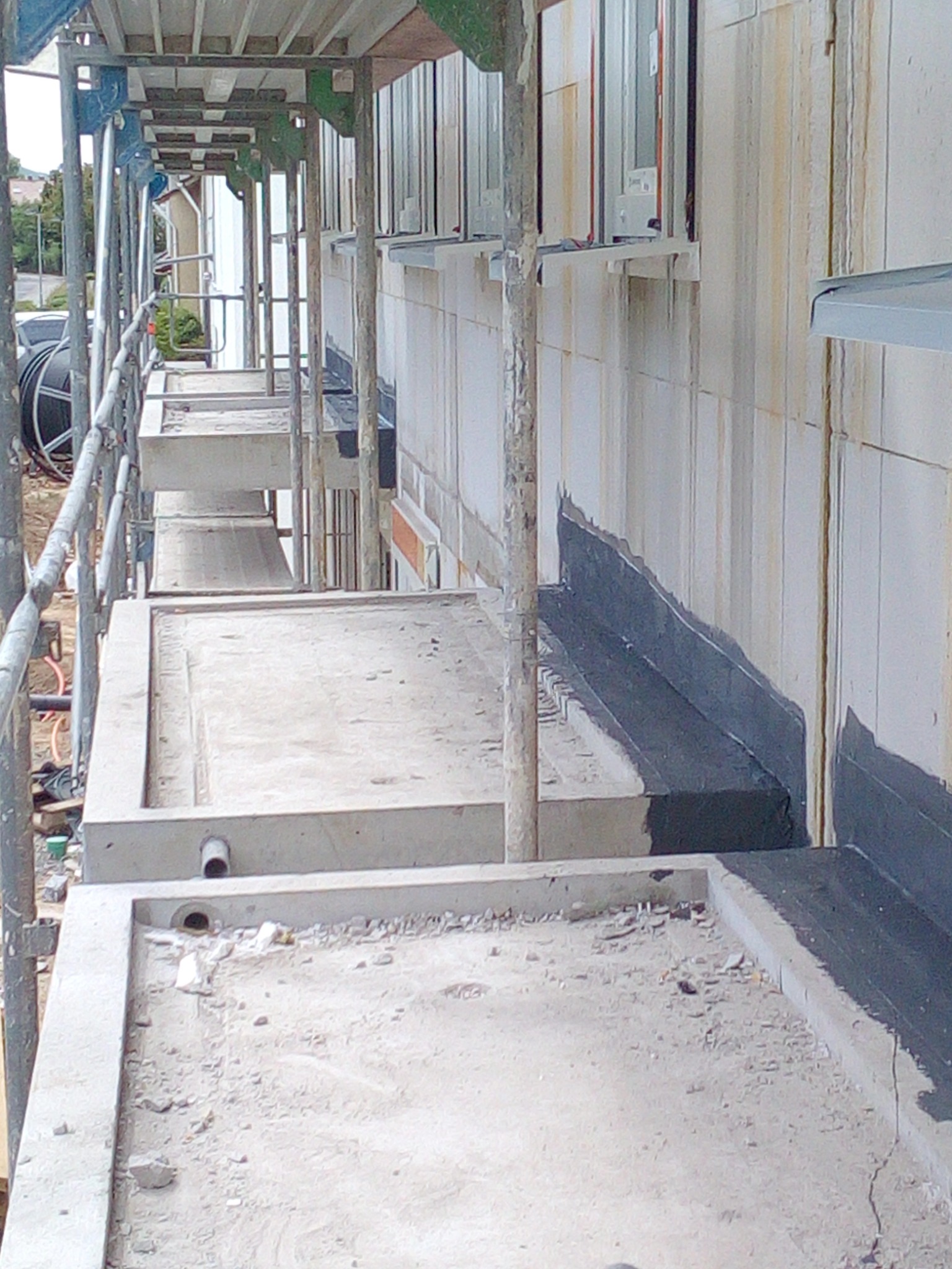 Ściana budynku z widoczną izolacją przeciwwilgociową na fundamencie, wykonana z białych bloczków, z widocznymi śladami zabrudzeń i zacieków, oraz ustawione rusztowanie.