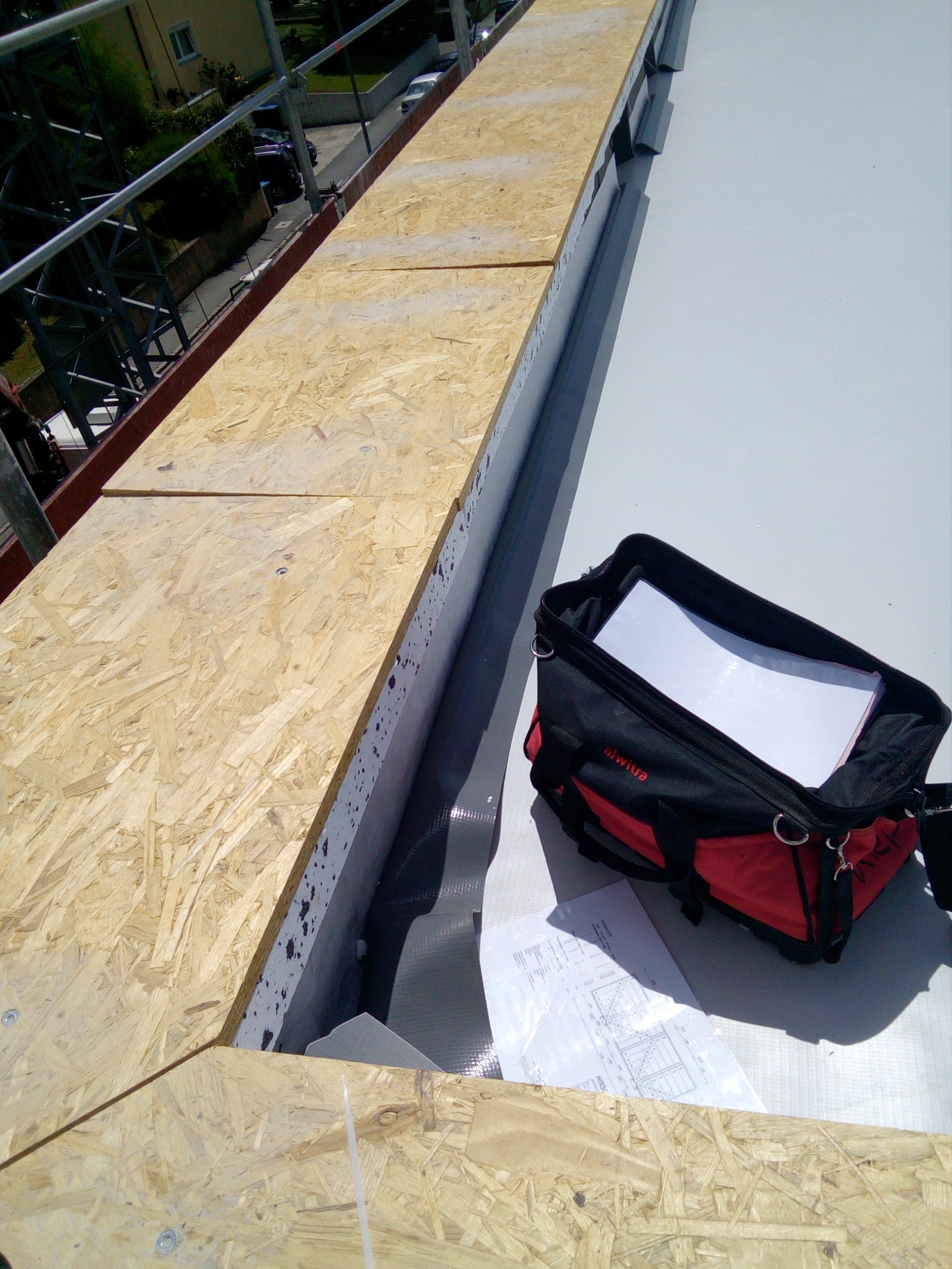 Widok z góry na fragment dachu w trakcie budowy: płyty OSB ułożone obok membrany dachowej, torba narzędziowa i plany na białym podłożu, rusztowanie w tle.