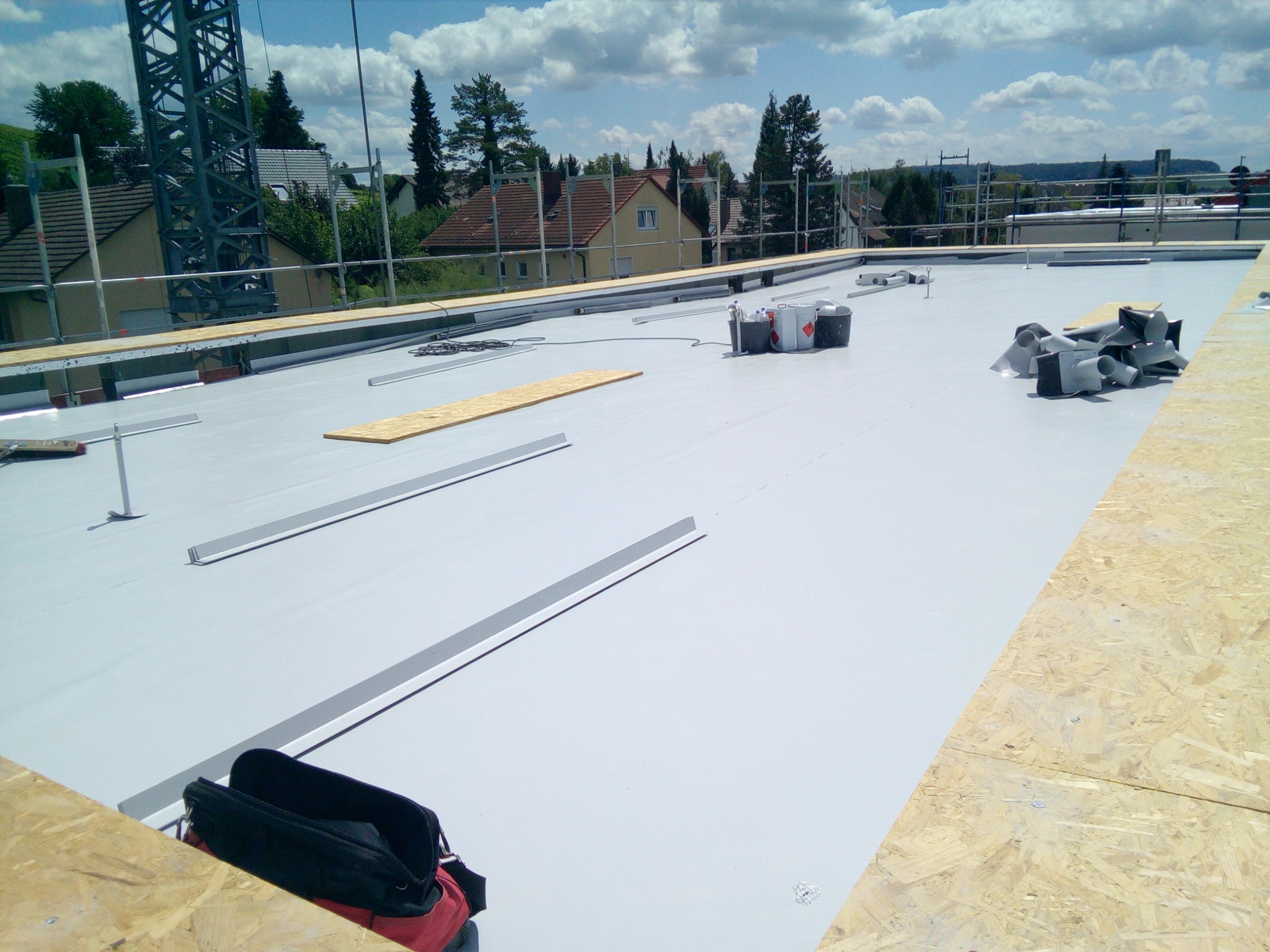 Płaski dach pokryty jasną membraną hydroizolacyjną z widocznymi elementami montażowymi, narzędziami i wentylacją, przygotowany do dalszych prac dekarskich, z widokiem na okoliczną zabudowę...