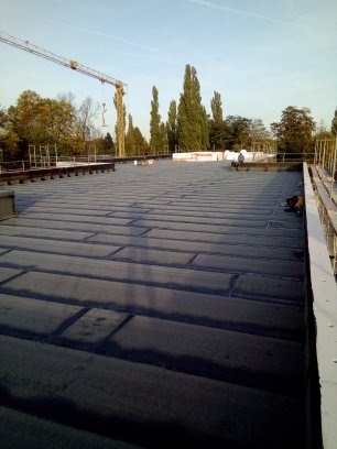 Płaski dach pokryty papą termozgrzewalną, widoczne zgrzewy, w tle dźwig budowlany i drzewa.