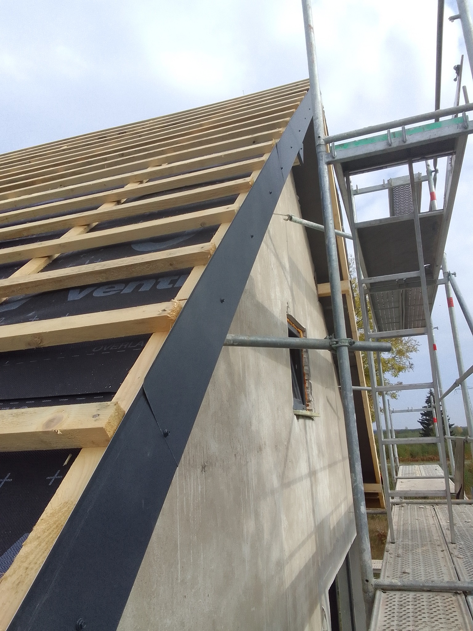 Budowa dachu: widok na drewnianą konstrukcję więźby pokrytą czarną membraną dachową, z zamontowaną obróbką blacharską przy krawędzi dachu i rusztowaniem w tle.